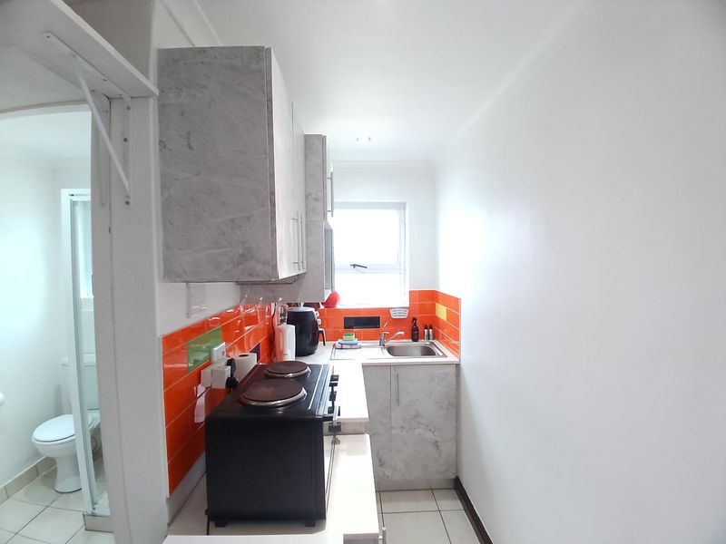 Compact kitchen featuring modern grey cabinetry, vibrant orange tiled splashback, stainless steel sink, and tiled flooring, with an adjacent bathroom visible.