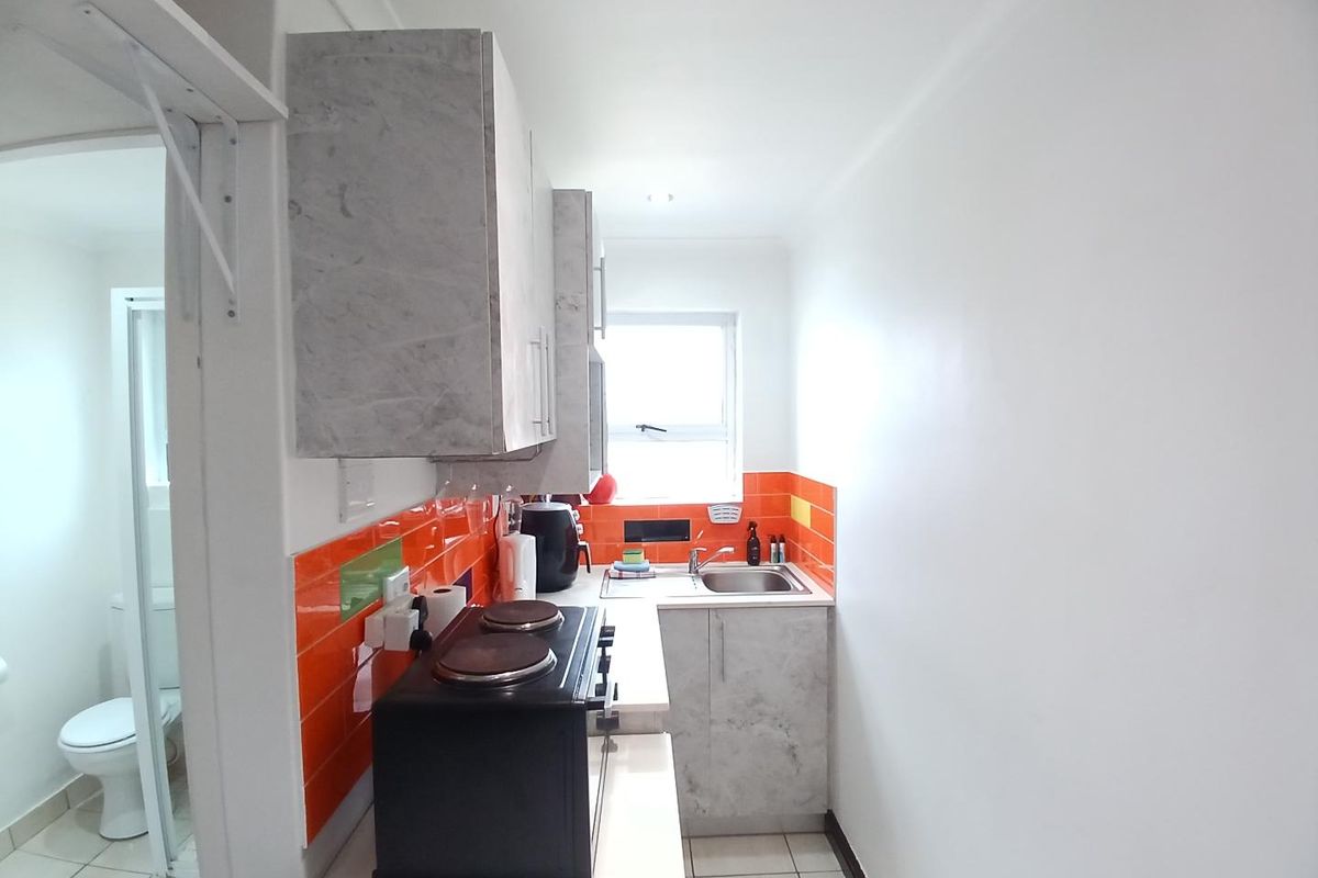 Compact kitchen featuring modern grey cabinetry, vibrant orange tiled splashback, stainless steel sink, and tiled flooring, with an adjacent bathroom visible.