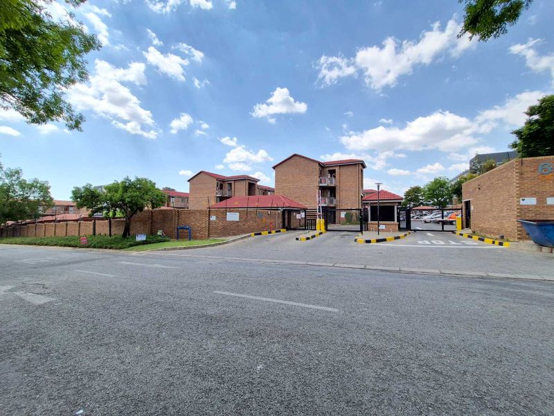 Street view of the apartment complex entrance featuring a brick boundary wall, security gate, and guard house.