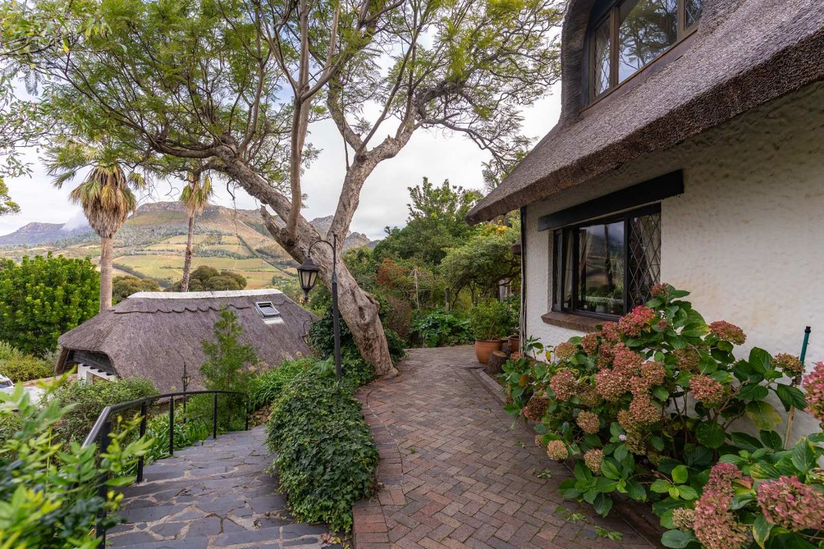A charming exterior view featuring a thatched roof cottage, brick pathway, and landscaped garden with scenic mountain views in the background.
