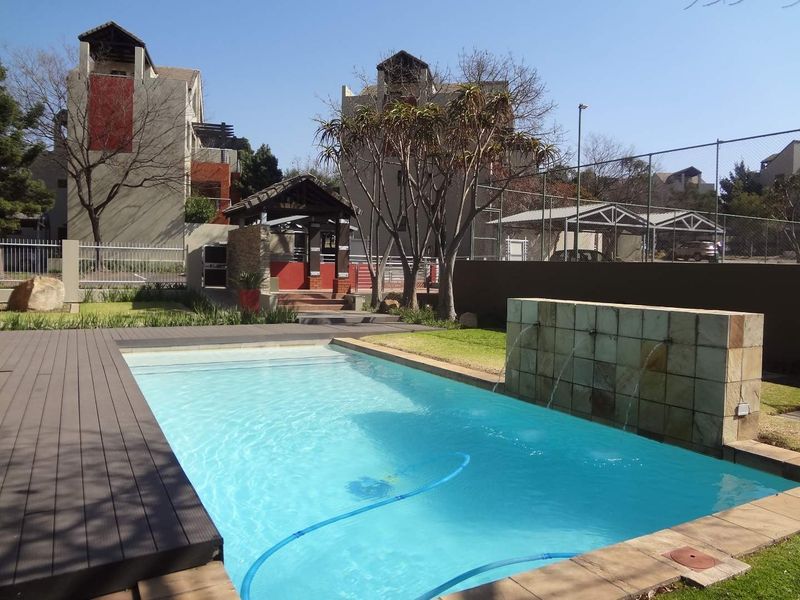 A well-maintained communal swimming pool area featuring a stone water feature, timber decking, and landscaped surroundings within an apartment complex.