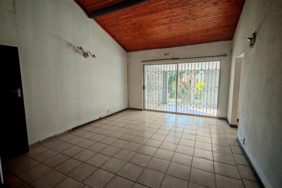 An empty living room featuring a vaulted timber-clad ceiling, tiled flooring, and sliding glass doors with security gates leading to the outdoor area.