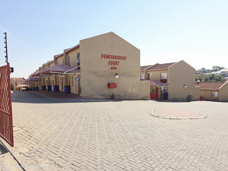 Exterior view of Peneyambeko Court apartment complex featuring paved driveway, metal security gate, electric fencing, and covered entrance awnings.