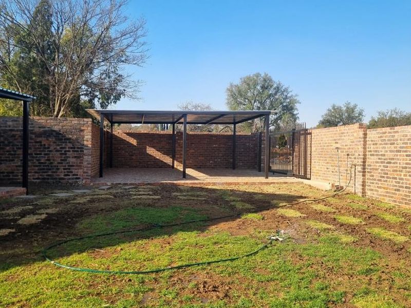A spacious rear yard featuring a metal carport structure, brick boundary walls, and a gated entrance with a lawn area.