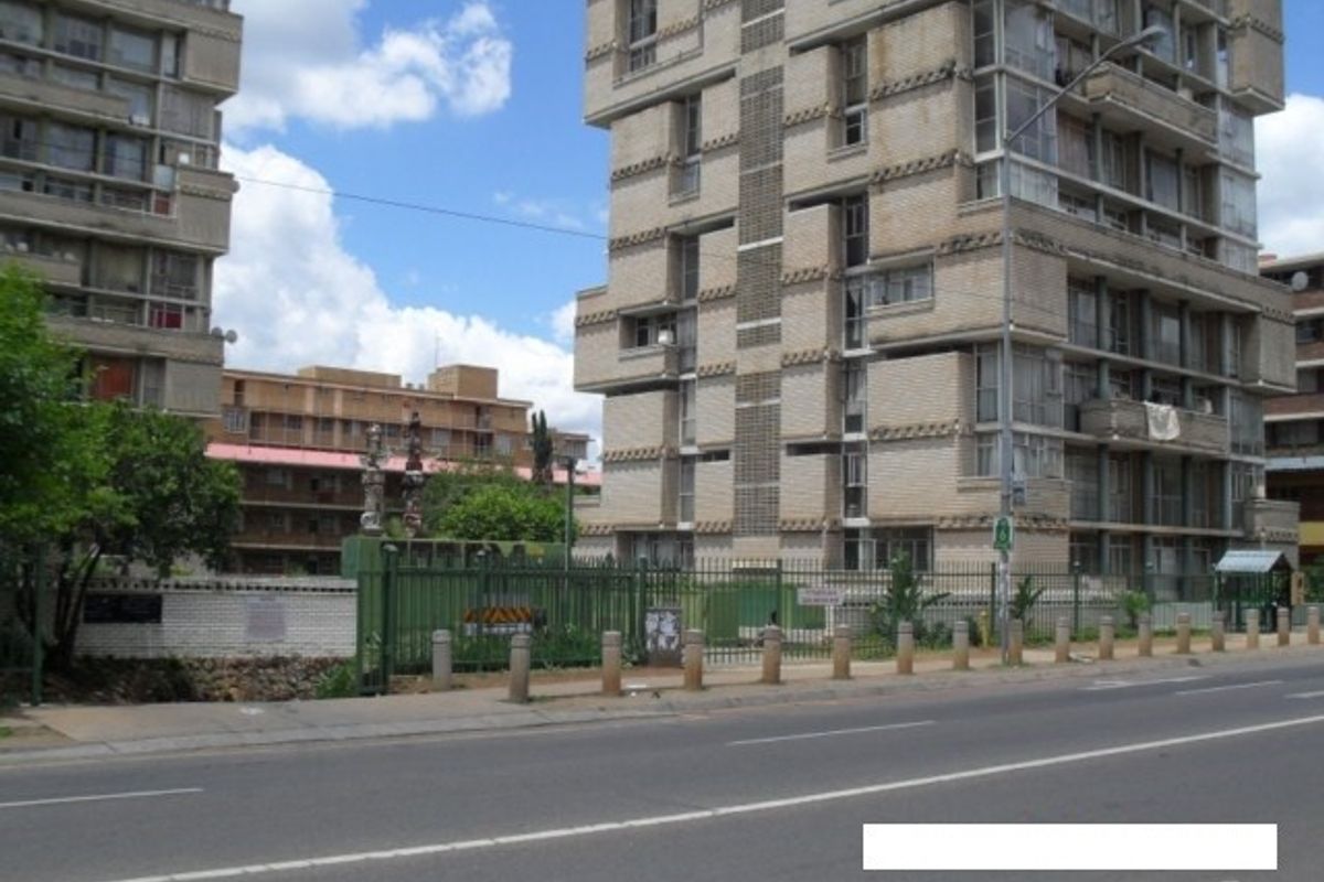 Street-level view of a multi-story apartment complex featuring brick facade construction and secure perimeter fencing along the roadside.