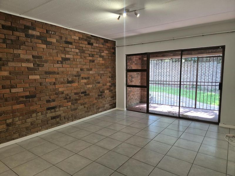 An empty living room featuring a prominent exposed brick wall, neutral ceramic floor tiles, and large sliding glass doors leading to an outdoor area.