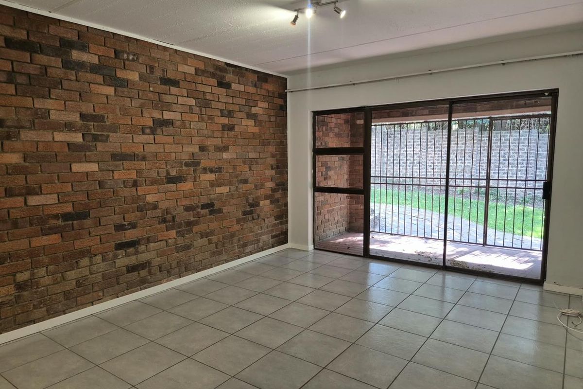 An empty living room featuring a prominent exposed brick wall, neutral ceramic floor tiles, and large sliding glass doors leading to an outdoor area.
