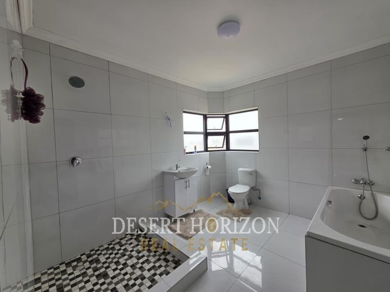 A spacious, fully tiled bathroom featuring a walk-in shower, a separate bathtub, a vanity basin, and a toilet with natural light from a corner window.