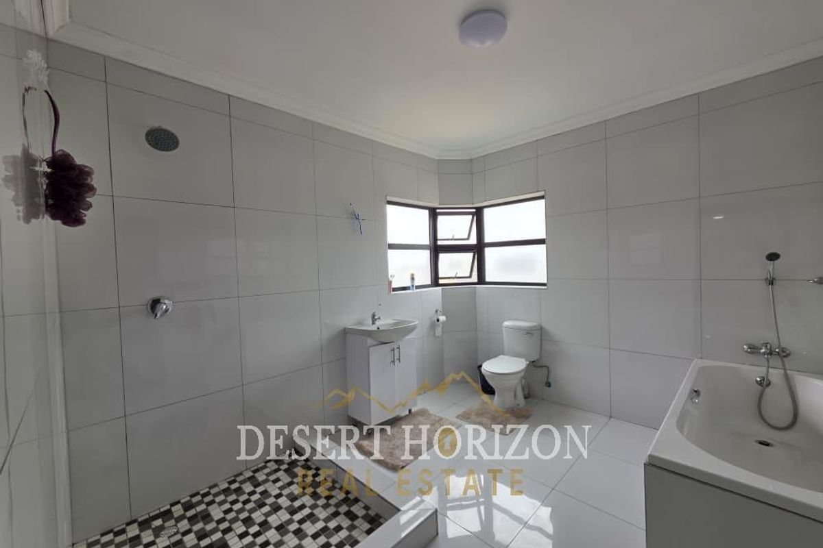 A spacious, fully tiled bathroom featuring a walk-in shower, a separate bathtub, a vanity basin, and a toilet with natural light from a corner window.