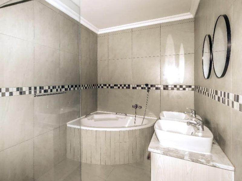 Modern bathroom featuring a corner bathtub, glass shower screen, and a double vanity with contemporary ceramic tiling and a decorative mosaic border.