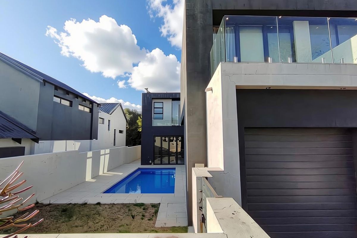 Modern multi-story home exterior featuring a swimming pool, paved patio area, and a glass-railed balcony.
