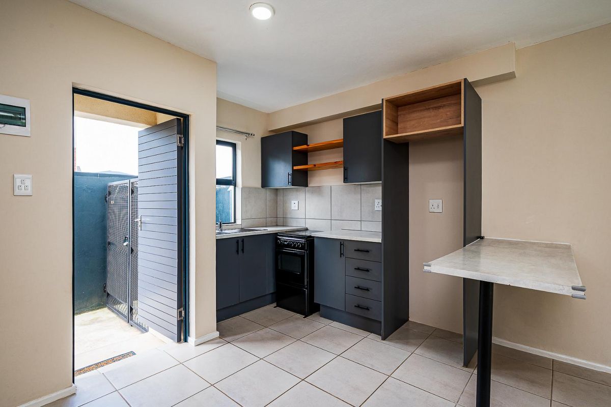 A compact kitchen featuring dark grey cabinetry, tiled splashbacks, a built-in stove, and a breakfast counter, with direct access to an outdoor area.