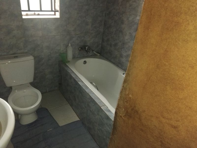 A compact bathroom featuring a built-in bathtub, toilet, and full-height wall tiling. The space requires cosmetic updates to improve its overall condition.