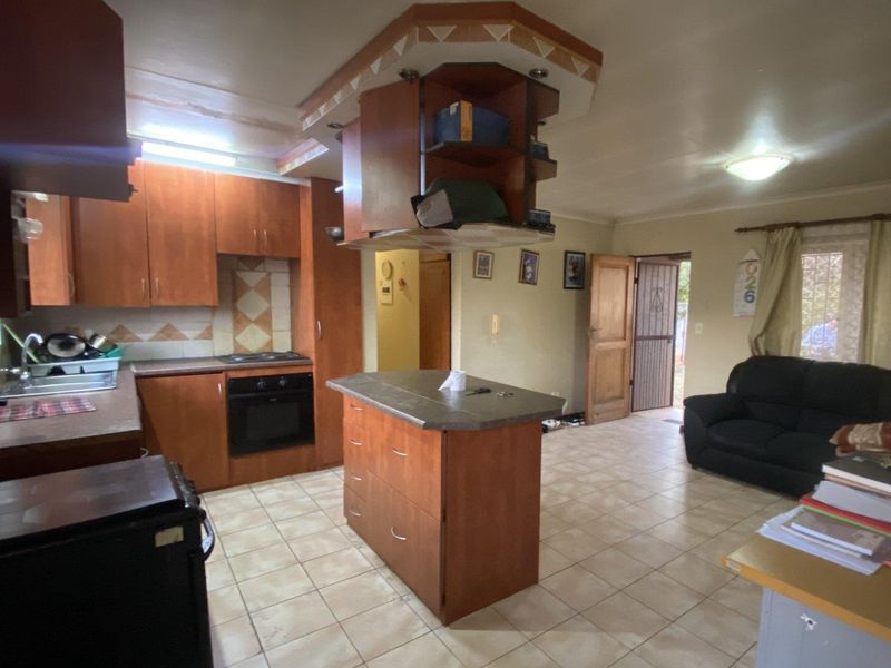 Open-plan kitchen and living area featuring wooden cabinetry, a central kitchen island, built-in oven, and tiled flooring throughout.