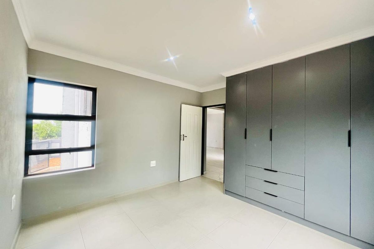 A bright, modern bedroom featuring neutral tiled flooring, recessed ceiling lighting, and extensive built-in grey cabinetry for ample storage.