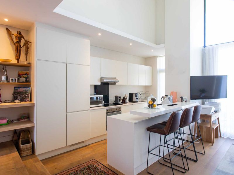 Modern open-plan kitchen featuring sleek white cabinetry, integrated appliances, and a breakfast bar, seamlessly flowing into the living area with high ceilings.