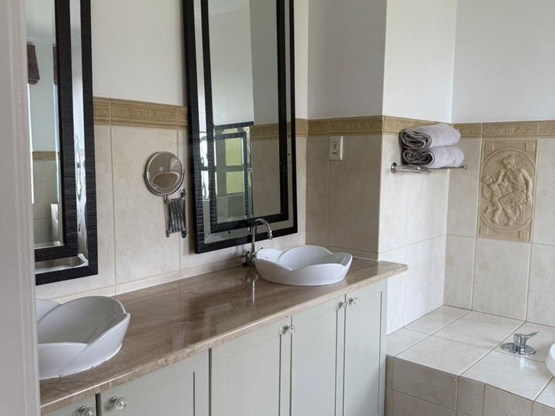 A well-maintained bathroom featuring a double vanity with vessel sinks, stone countertop, built-in cabinetry, and a tiled bathtub surround with decorative wall accents.