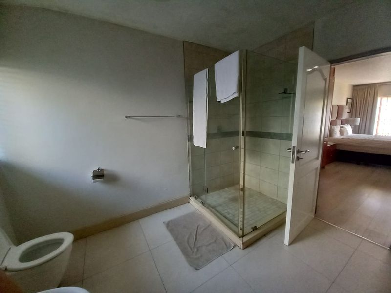 A functional bathroom featuring a glass-enclosed shower, wall-mounted toilet, and neutral floor tiling, with direct access to the adjacent bedroom.