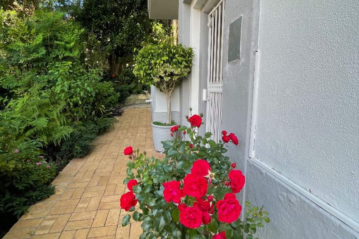 A well-maintained paved walkway along the side of the apartment building, featuring a secure entrance gate and lush garden surroundings.
