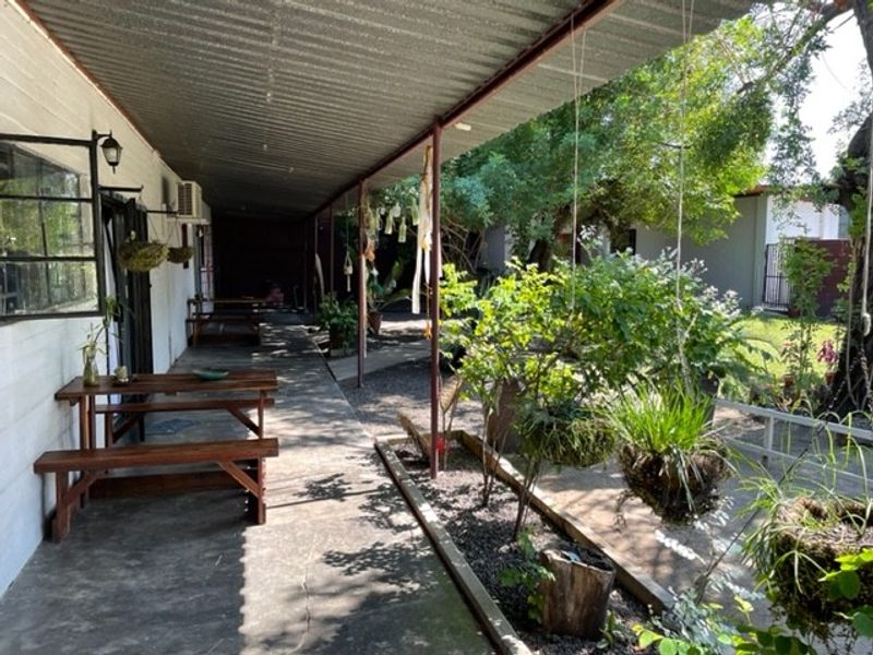 A covered outdoor patio area featuring a corrugated metal roof, concrete flooring, and metal support columns, overlooking a garden space.