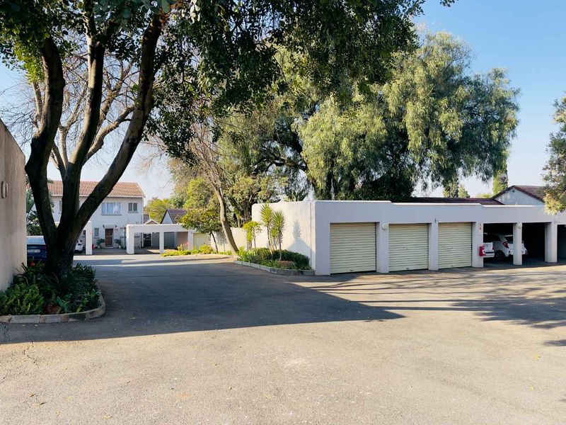 A view of the apartment complex exterior featuring a paved driveway, mature trees, and a row of secure garage units with roller shutter doors.