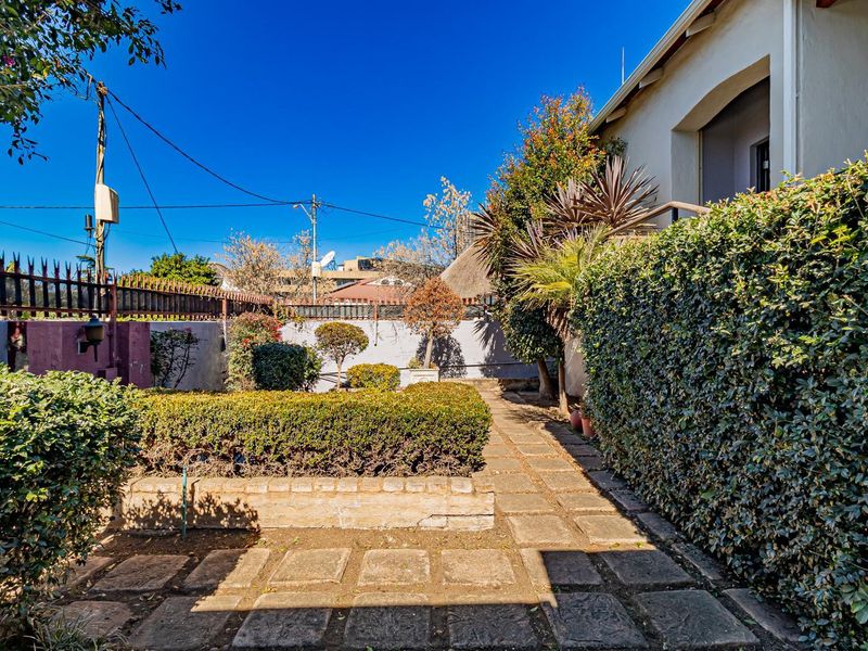 A rear garden area featuring a paved walkway, established hedging, and a brick retaining wall along the property boundary.