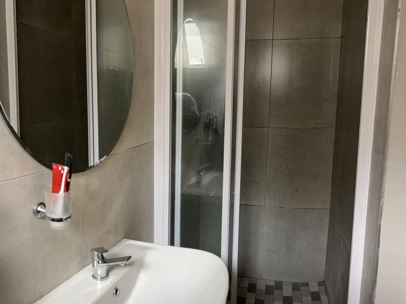 A compact bathroom featuring a pedestal basin with a chrome mixer tap, a round wall mirror, and a tiled shower enclosure.