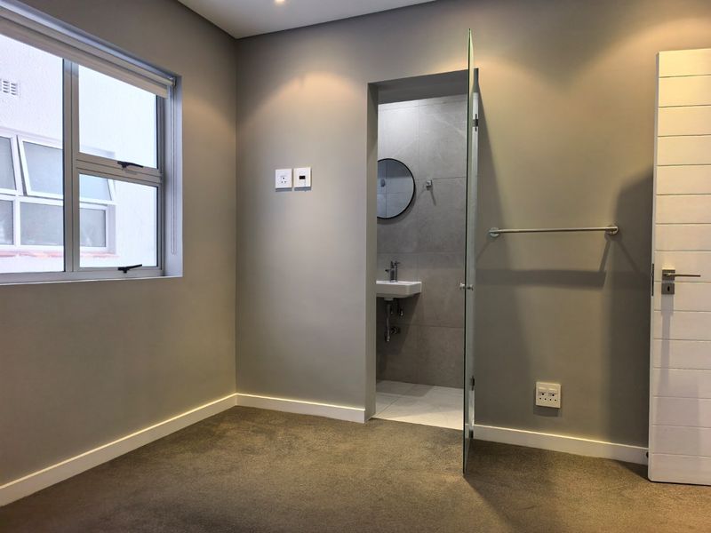 A clean, neutral-toned bedroom featuring plush carpeting, modern recessed lighting, and an integrated en-suite bathroom with contemporary finishes.