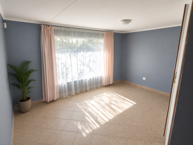 A bright, empty bedroom featuring neutral ceramic floor tiles, blue-toned walls, and a large window with curtains, offering a clean and versatile living space.