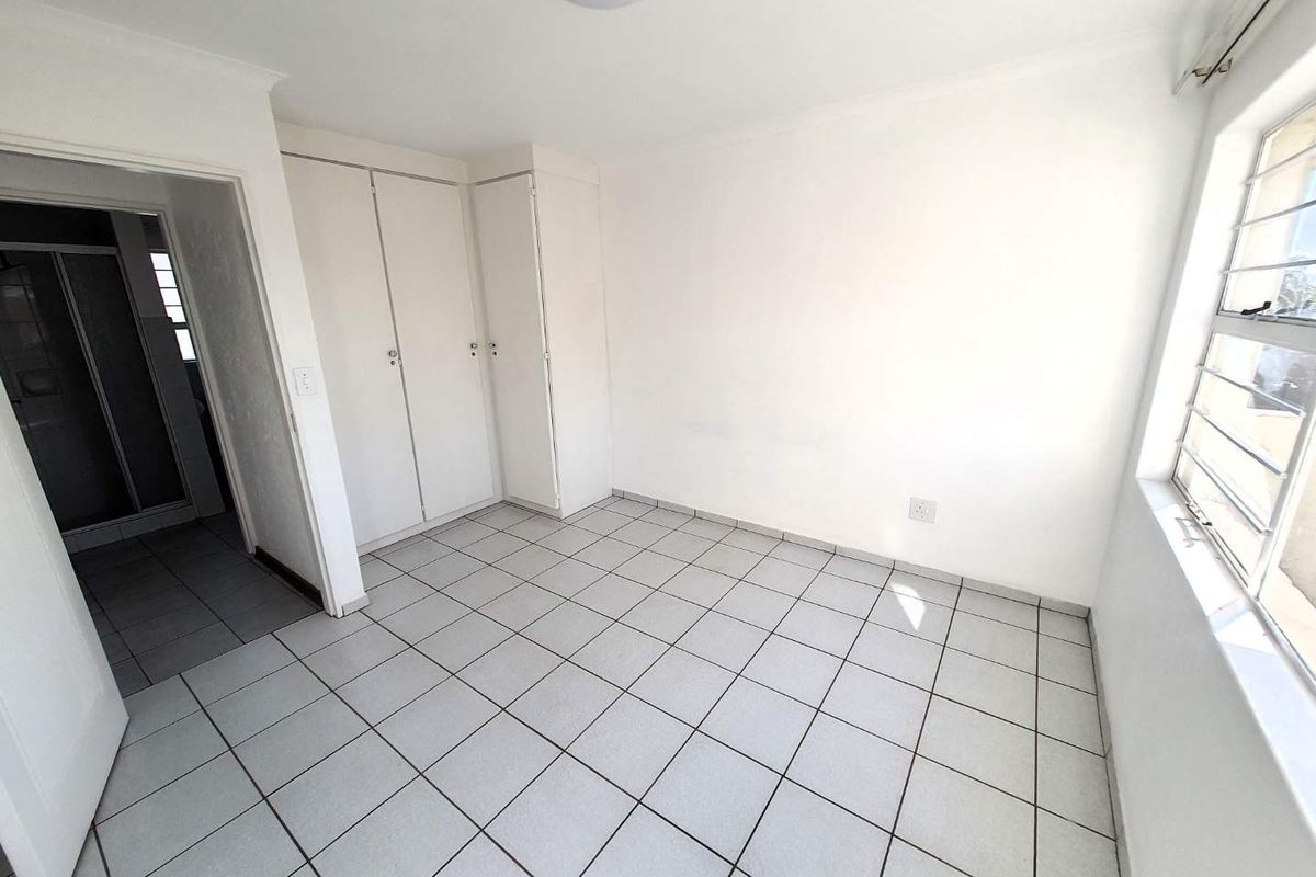 A bright, empty bedroom featuring tiled flooring, built-in cupboards, and a window with security bars, leading to an adjacent bathroom.