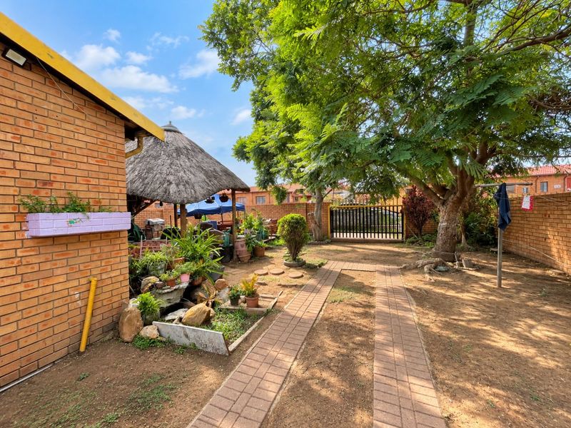 A rear garden area featuring a brick boundary wall, a thatched lapa, and a paved walkway leading to a metal gate.