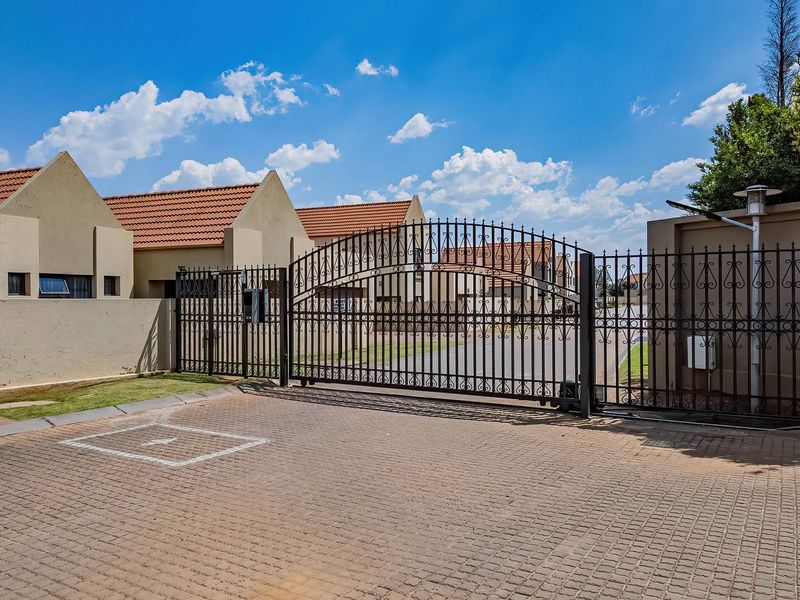 A secure gated entrance to a residential cluster complex featuring a paved driveway, perimeter walling, and wrought iron security gates.