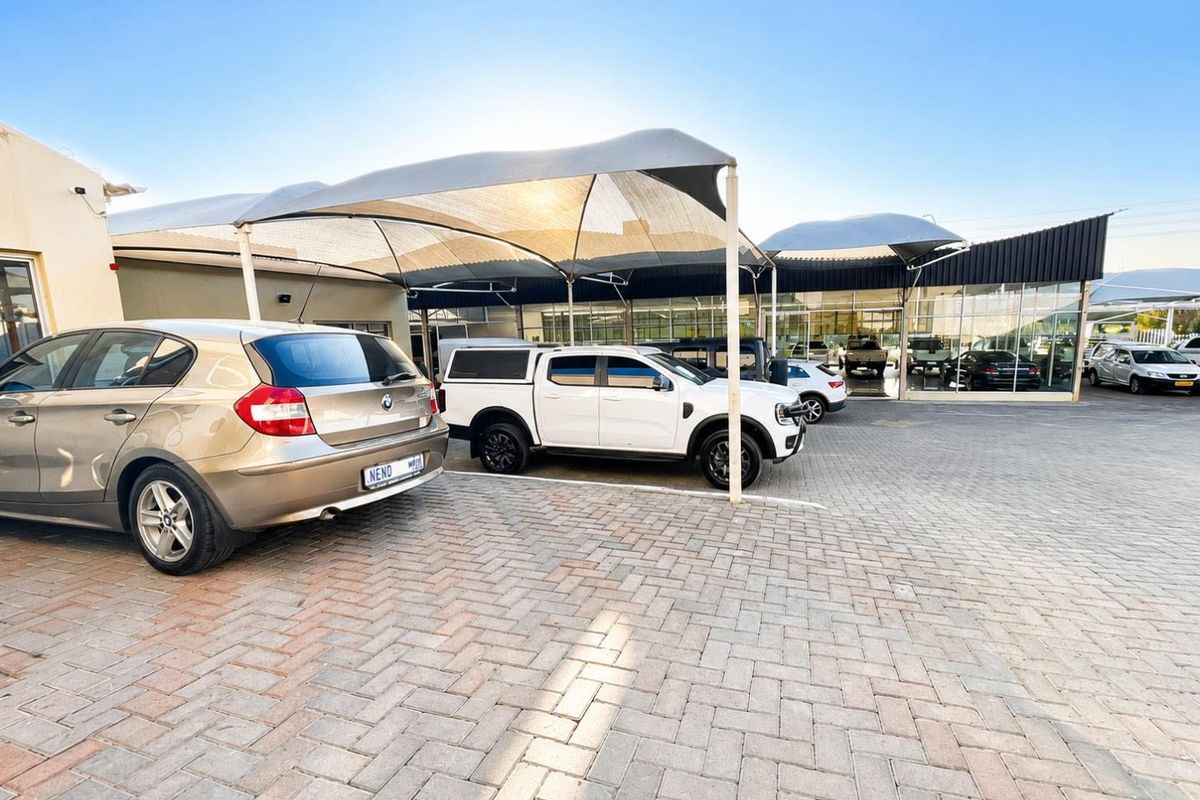Exterior view of a retail property featuring a paved parking area with shade cloth structures and large glass storefront windows.