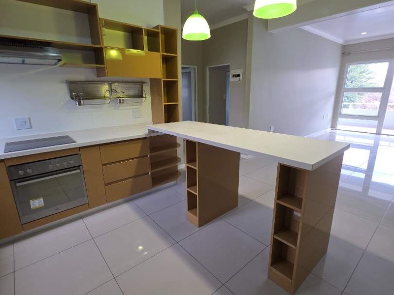 Modern open-plan kitchen featuring light wood cabinetry, a central island, integrated appliances, and polished floor tiles, flowing into a bright living area.