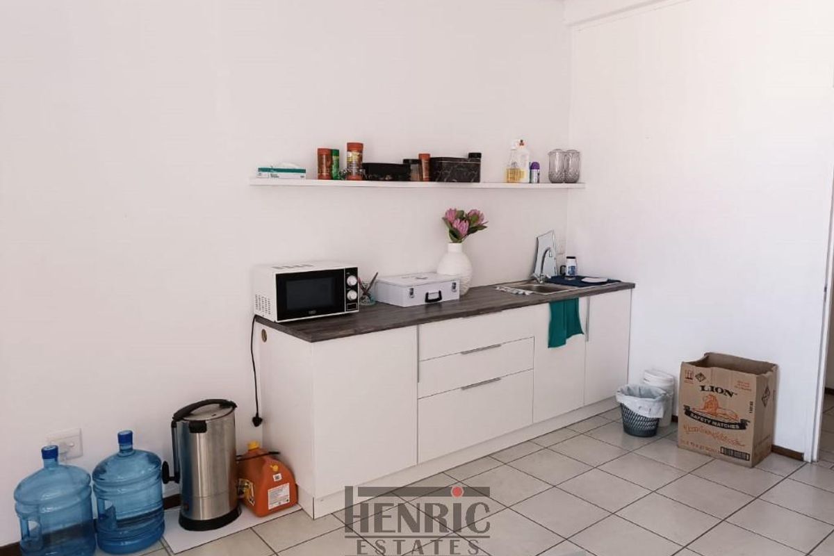 A compact office kitchenette featuring white cabinetry, a dark laminate countertop, a stainless steel sink, and a floating wall shelf.
