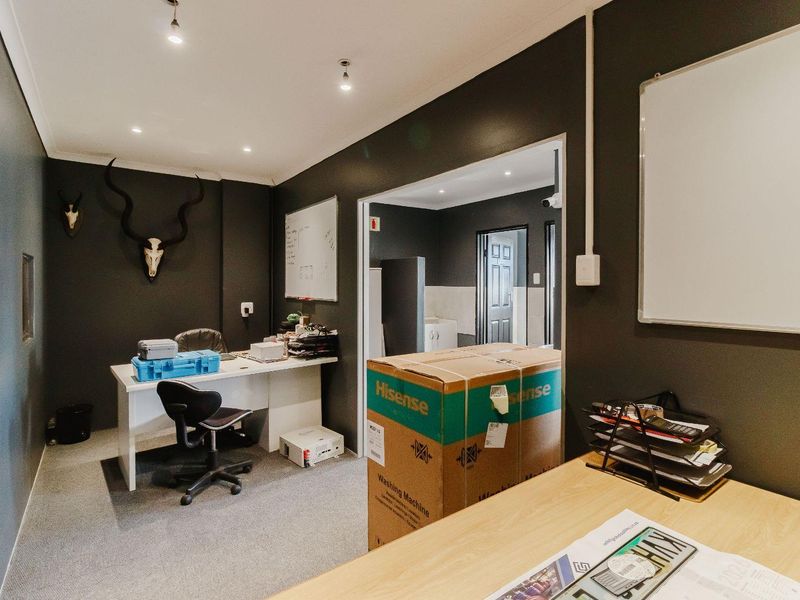 A functional office space featuring dark painted walls, recessed lighting, and carpeted flooring, with an adjoining utility area visible through the doorway.