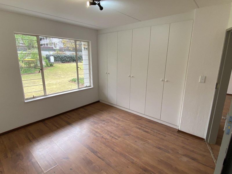 A bright, empty bedroom featuring laminate flooring, large windows with security bars, and extensive built-in cupboard storage.