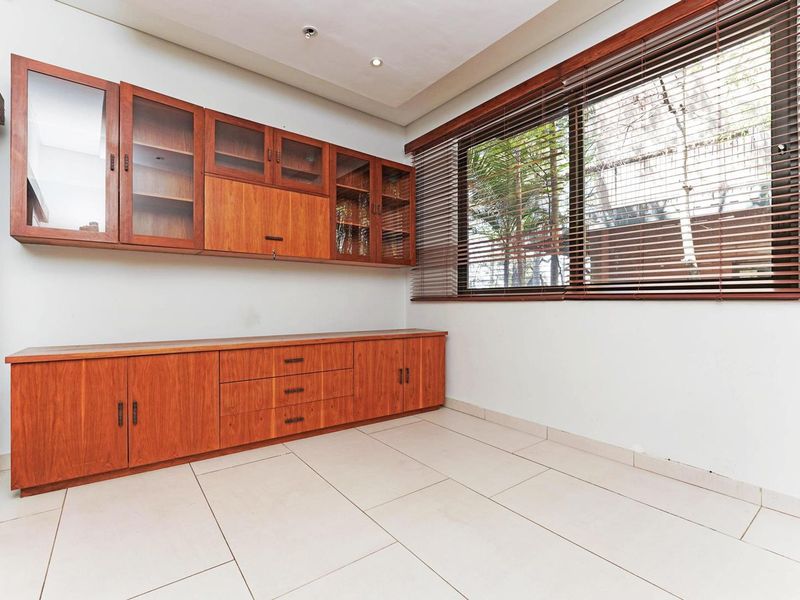 A bright, empty office space featuring extensive built-in wooden cabinetry, large windows with venetian blinds, and clean tiled flooring.