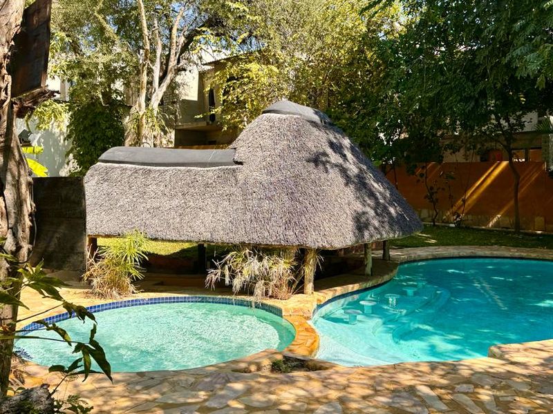 A tranquil outdoor pool area featuring a unique thatched-roof gazebo with a swim-up bar, surrounded by lush trees and natural stone paving.