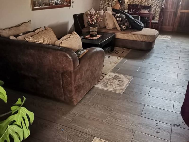 A living room featuring dark wood-look tiled flooring with decorative inset tiles, a large wall mirror, and access to an adjacent room.