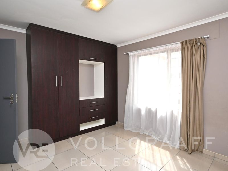 A bright, empty bedroom featuring dark wood built-in cupboards, neutral tiled flooring, and a large window with curtain tracks.