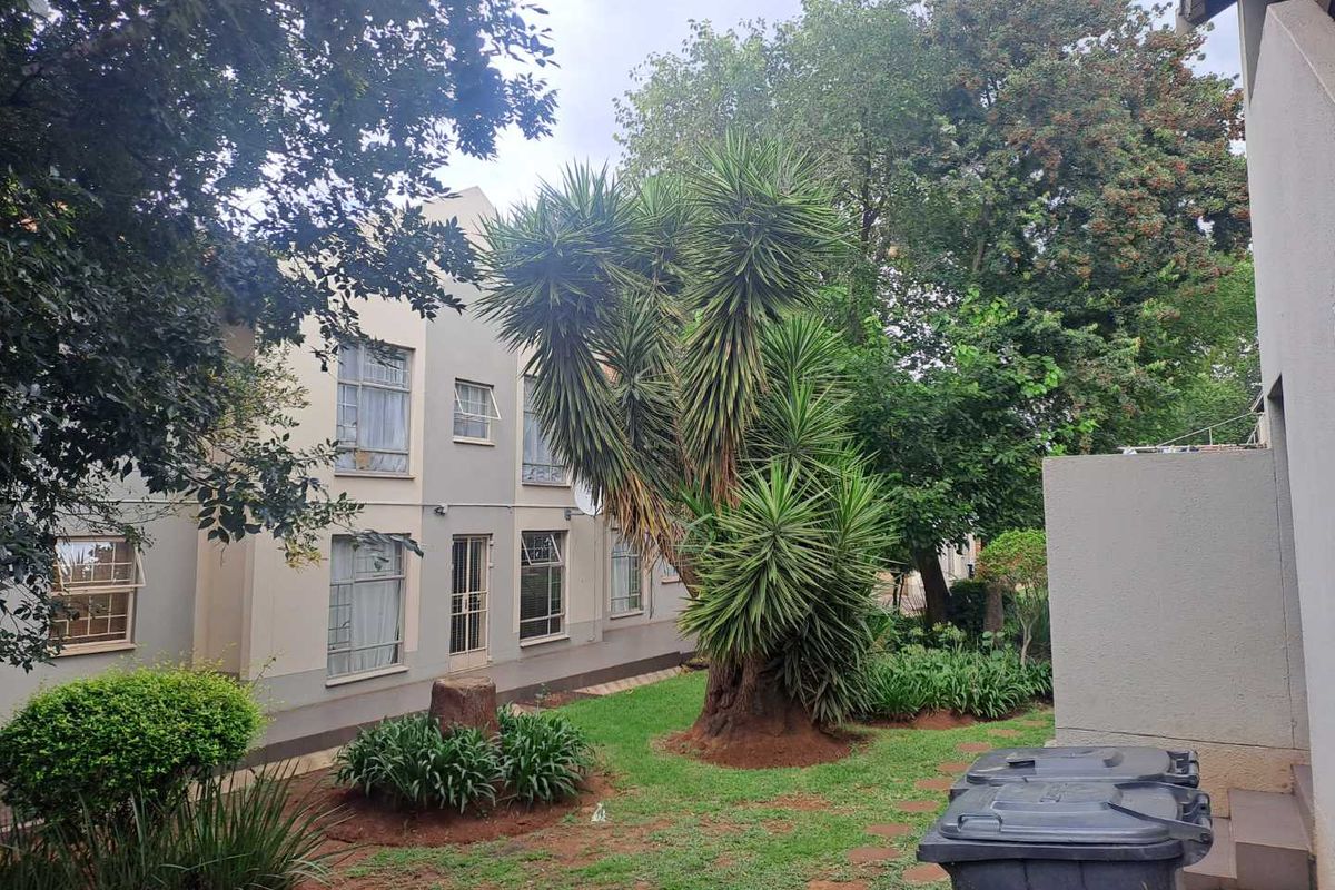 A view of the rear exterior of the apartment building, featuring a shared garden area with mature trees and a paved walkway.