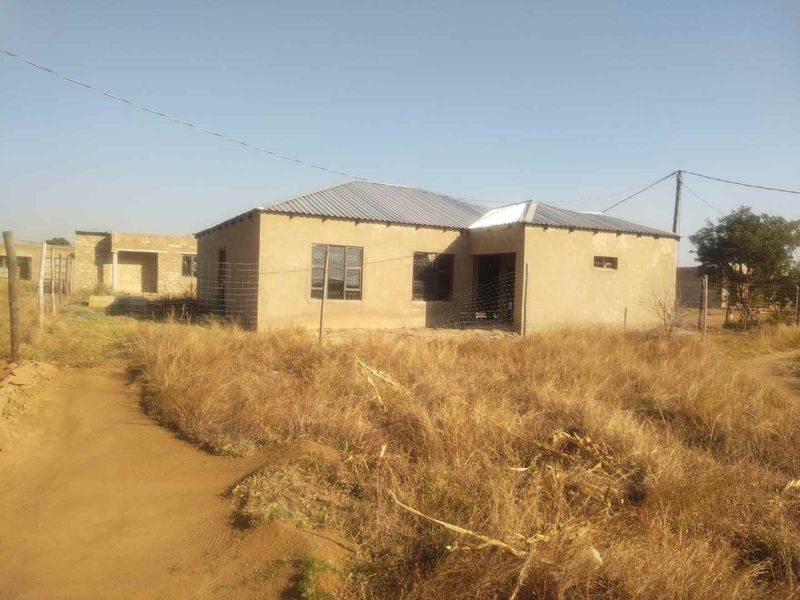 A single-story residential property under construction, featuring unpainted plastered walls, a corrugated metal roof, and steel window frames on a large plot.