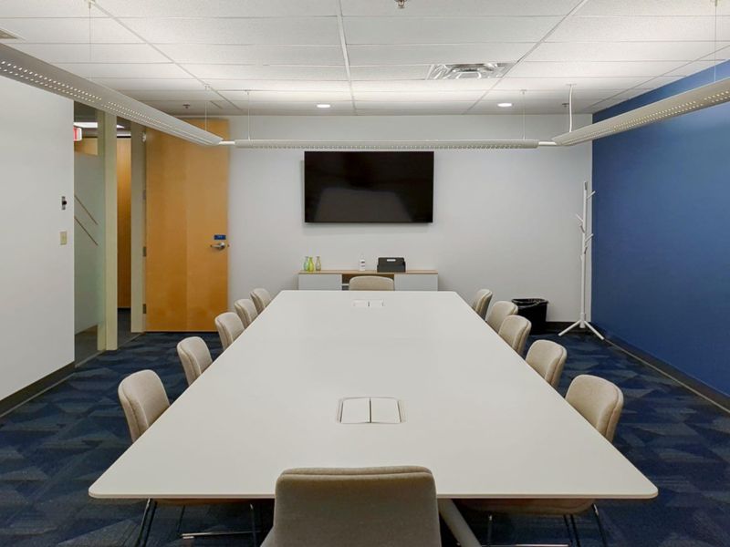 A professional office boardroom featuring a large central table, suspended lighting, and a wall-mounted display screen in a clean, modern workspace.
