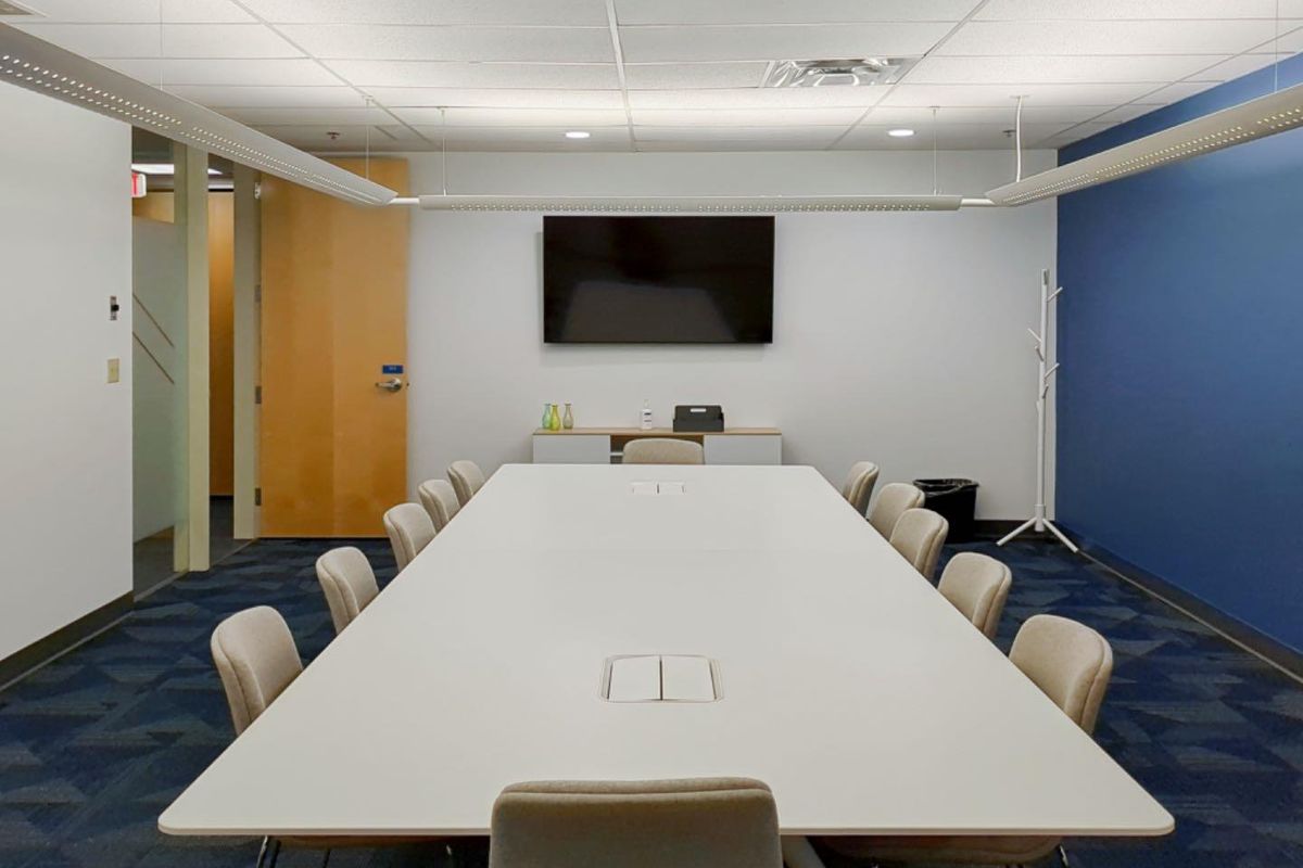 A professional office boardroom featuring a large central table, suspended lighting, and a wall-mounted display screen in a clean, modern workspace.