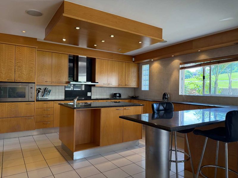 Spacious kitchen featuring warm wooden cabinetry, granite countertops, a central island with breakfast bar, and integrated appliances under a modern bulkhead ceiling.