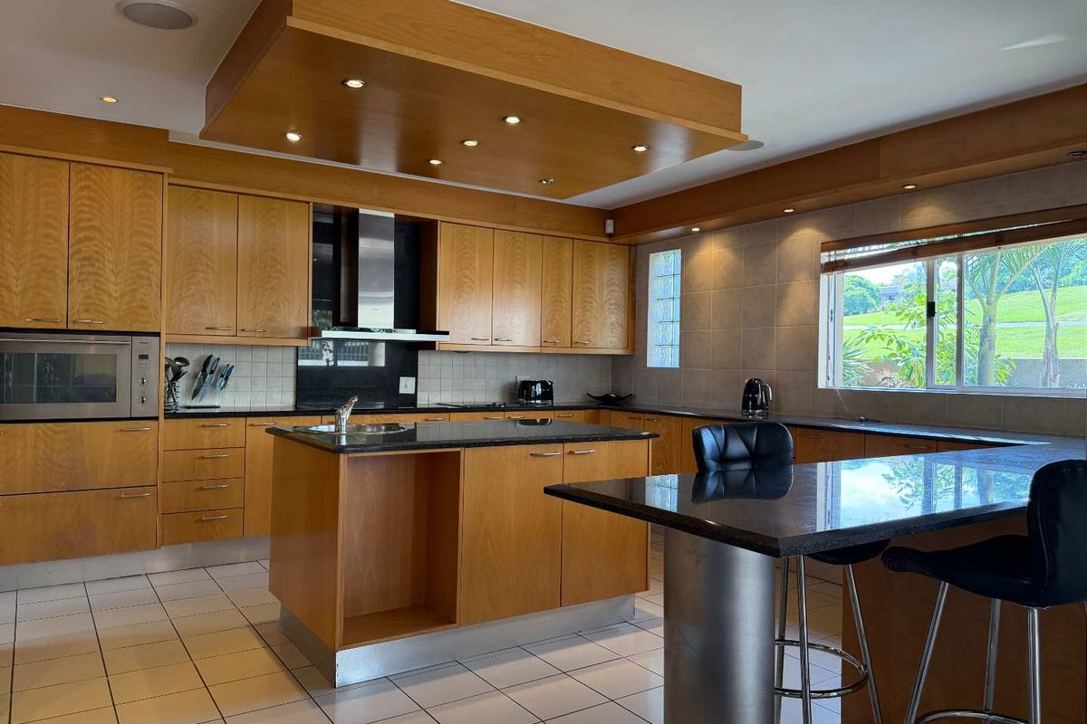 Spacious kitchen featuring warm wooden cabinetry, granite countertops, a central island with breakfast bar, and integrated appliances under a modern bulkhead ceiling.