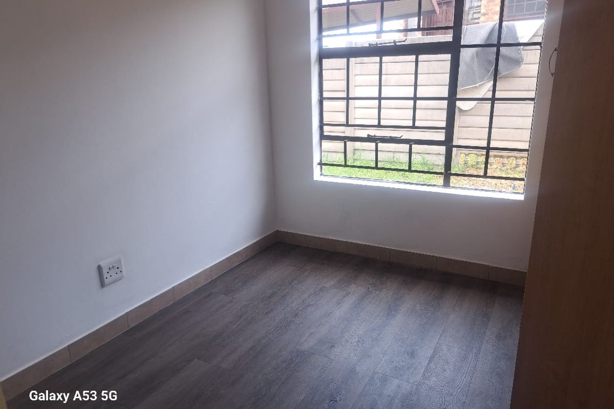 A clean, empty bedroom featuring modern wood-look laminate flooring, a large window with security bars, and a built-in cupboard.