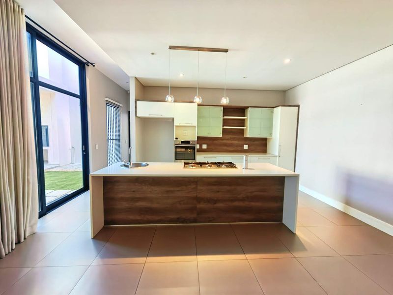 Modern open-plan kitchen featuring a central island with a gas hob, built-in oven, sleek cabinetry, and large glass doors leading to the garden.