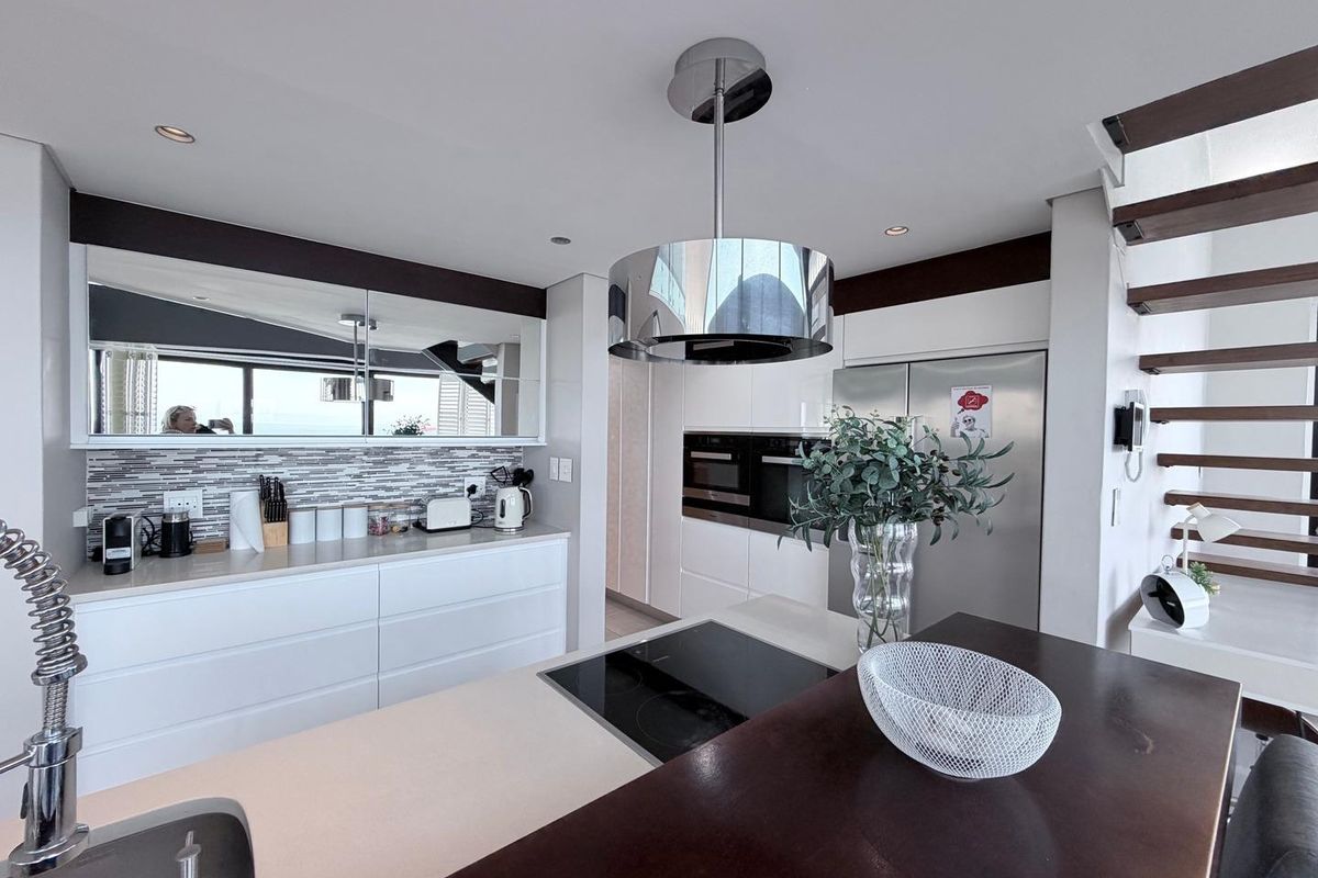 Modern kitchen featuring a central island with induction cooktop, sleek white cabinetry, integrated appliances, and a stylish overhead extractor fan, adjacent to a staircase.